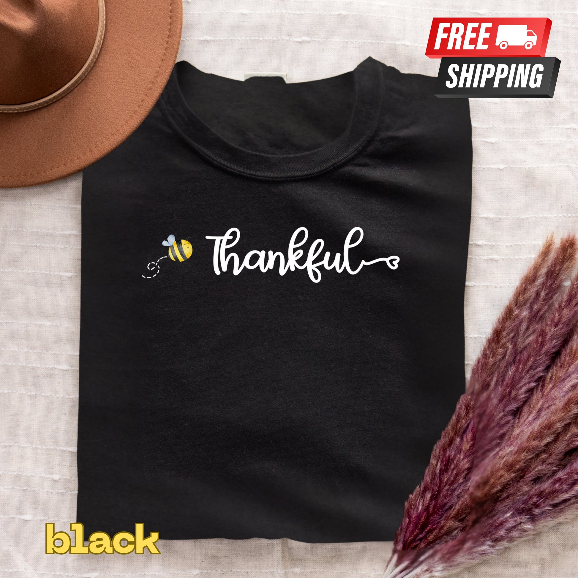 Black t-shirt with 'Thankful' design and bee icon, placed on a textured surface with a hat and dried plants.