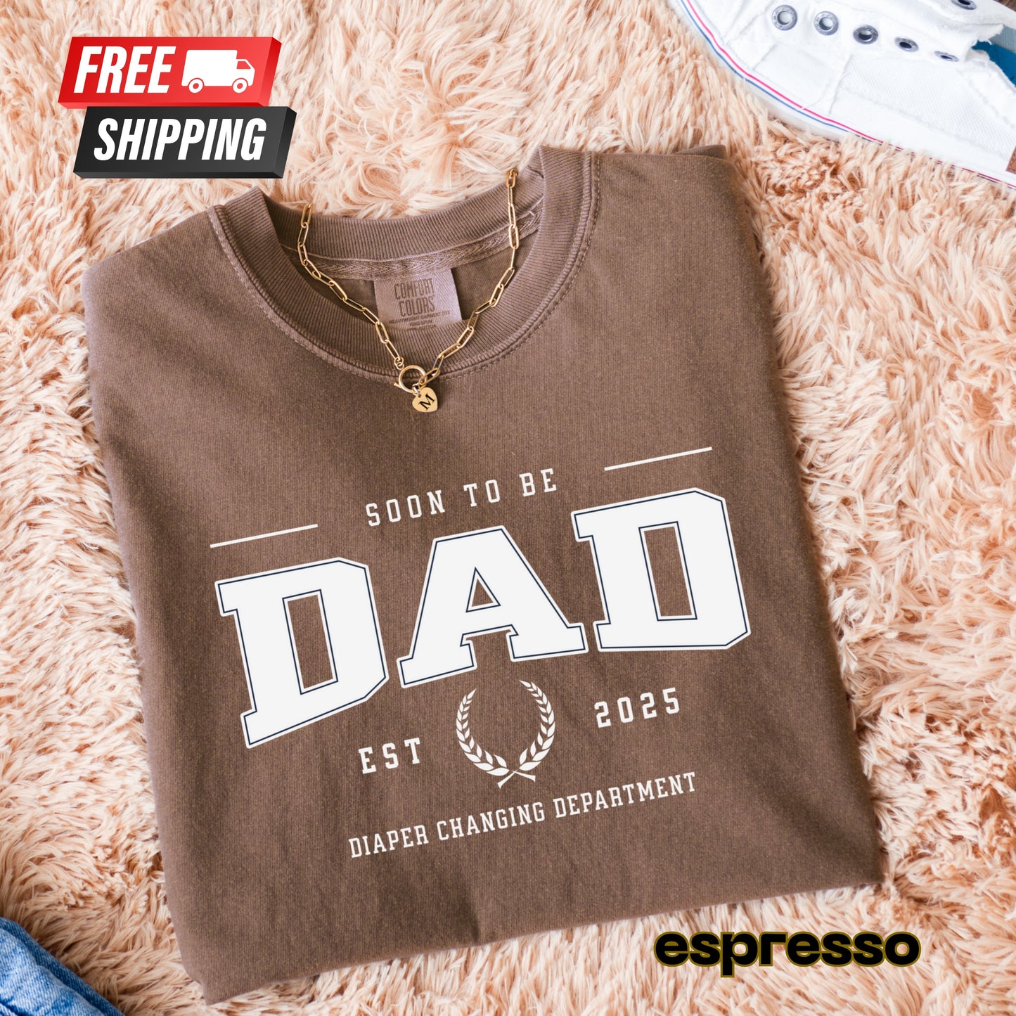 Folded brown sweatshirt with 'Soon to be Dad' text on a fluffy beige surface, featuring free shipping badge and espresso brand name.