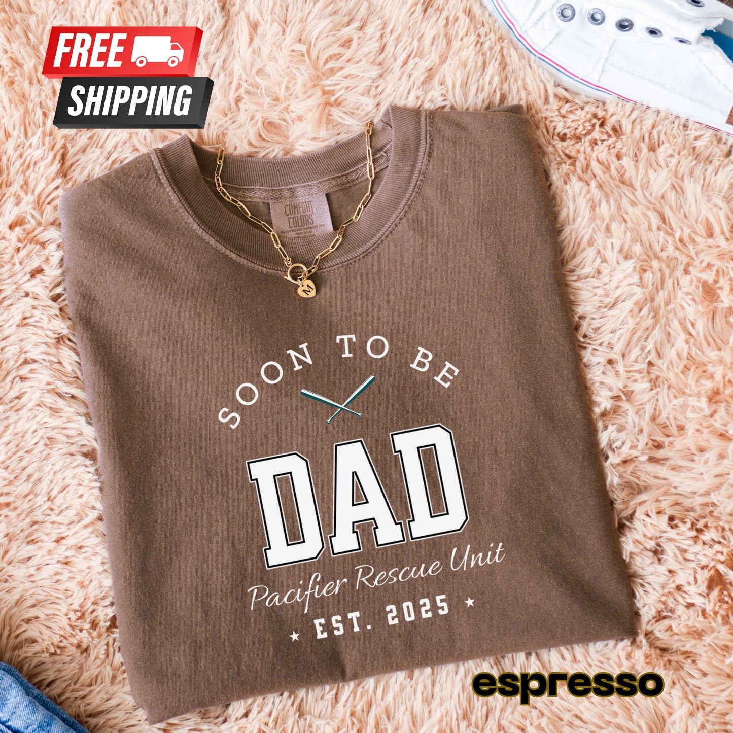 Folded brown sweatshirt with 'Soon to be Dad' text on a fluffy beige surface, featuring free shipping icon and espresso brand logo.