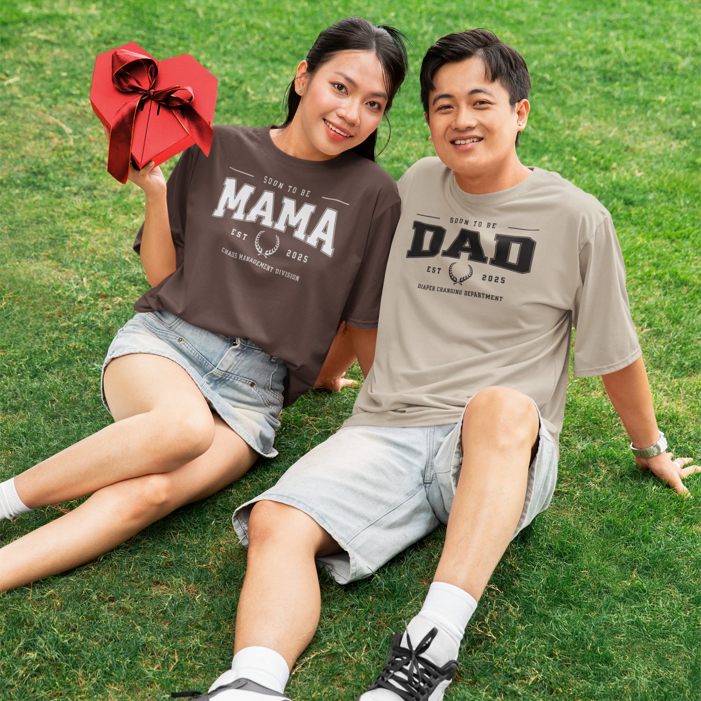 Two people sitting on grass wearing t-shirts with 'MAMA' and 'DAD' text, holding a red gift bag.