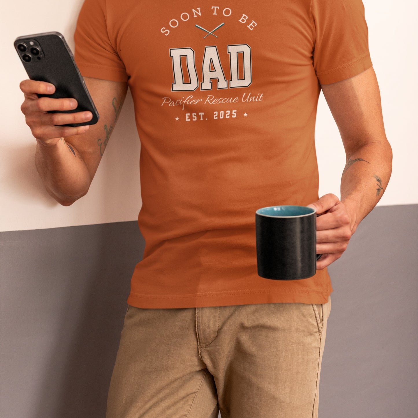 Person wearing an orange t-shirt with 'Soon to be Dad' text, holding a phone and mug.