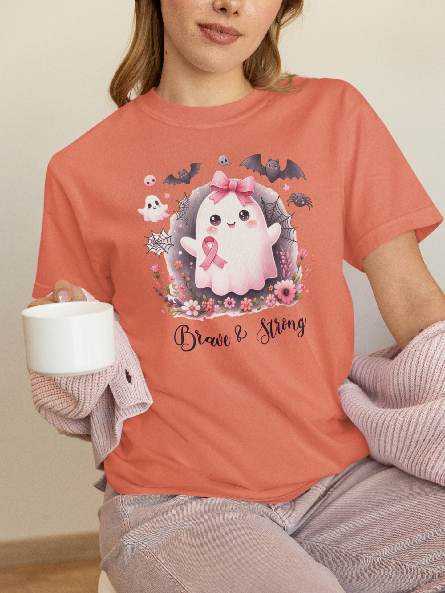 Person wearing a coral t-shirt with a ghost graphic and 'Brave & Strong' text, holding a white mug.