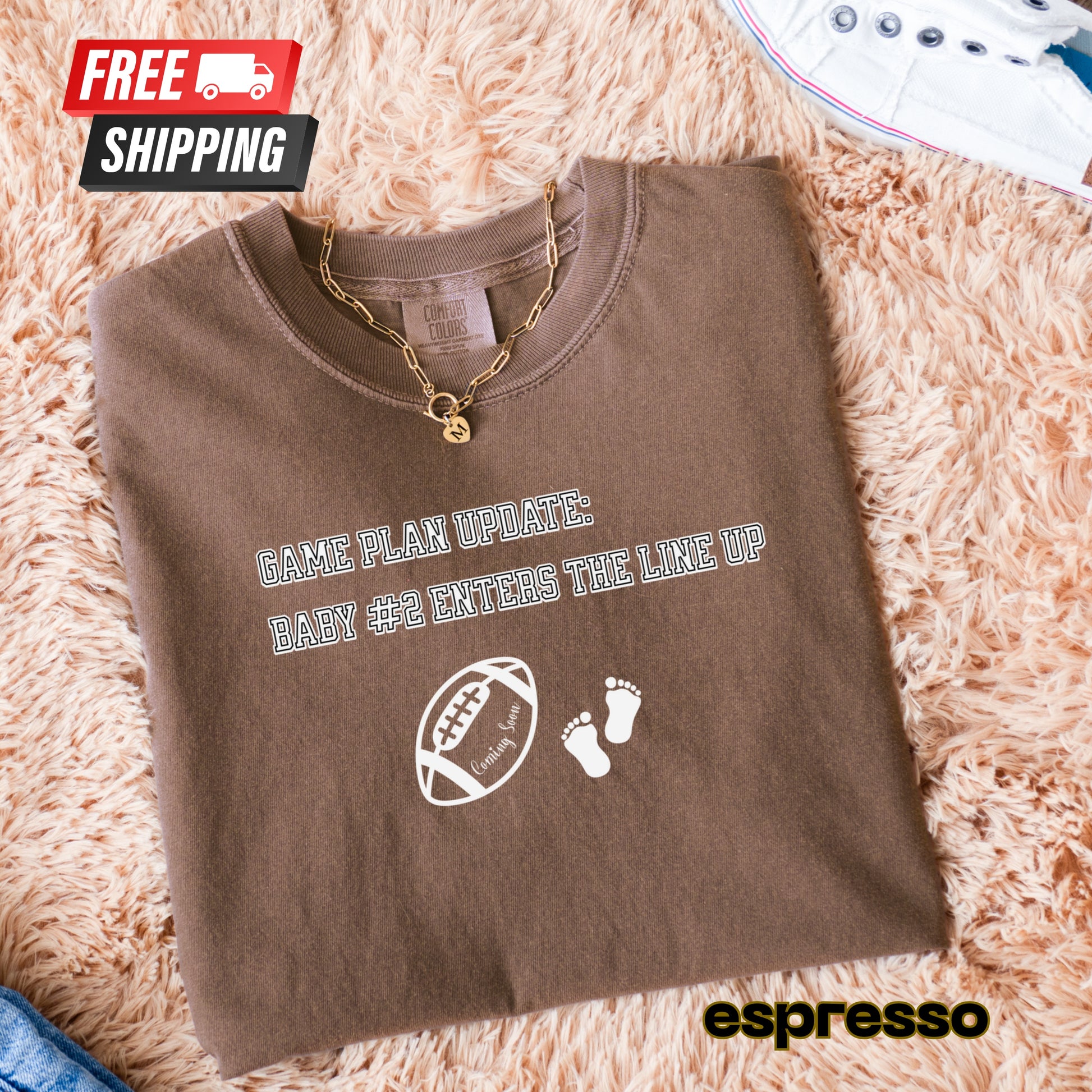 Folded brown t-shirt with baby announcement design on a fluffy surface, featuring free shipping icon and espresso brand.
