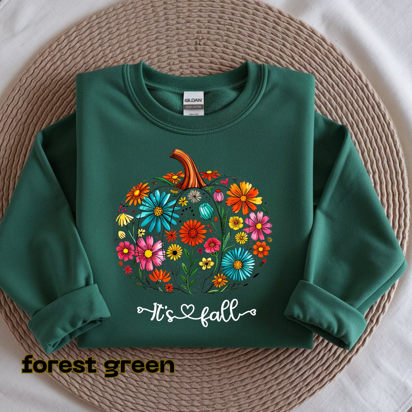 Green sweatshirt with floral pumpkin design and 'It's fall' text on a woven mat.