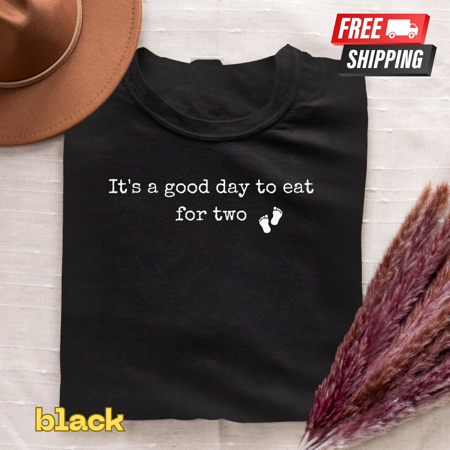 Black t-shirt with 'It's a good day to eat for two' text and footprints, free shipping label, and 'black' text on a textured surface.