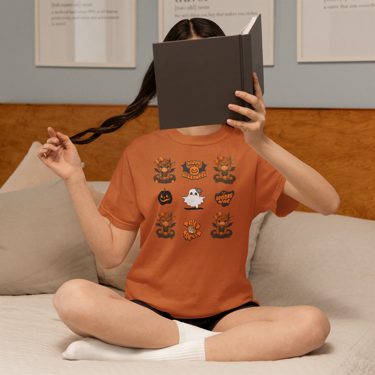 Person wearing an orange t-shirt with Halloween-themed designs, sitting on a couch and reading a book.