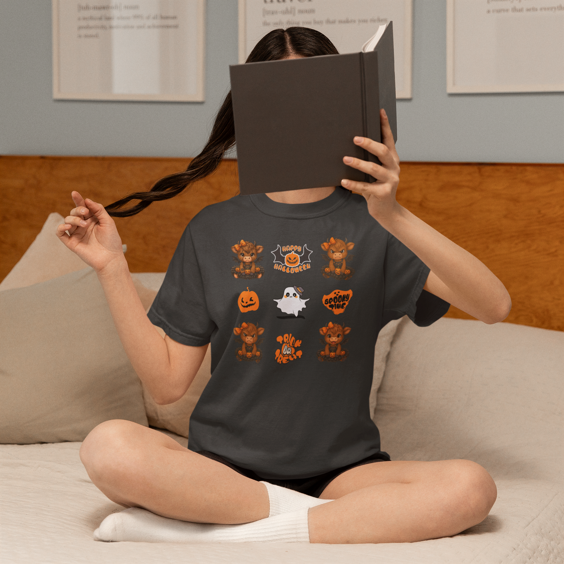 Person wearing a black t-shirt with Halloween-themed graphics, sitting on a couch and reading a book.