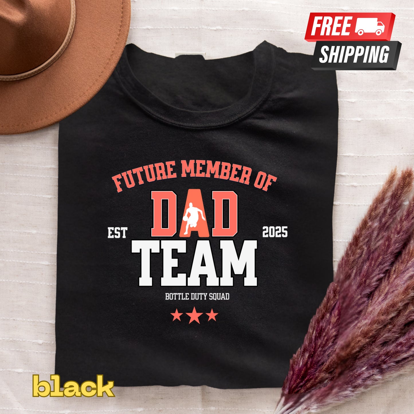 Black t-shirt with 'Future Member of Dad Team' text, free shipping label, and brown hat on a light wooden surface.