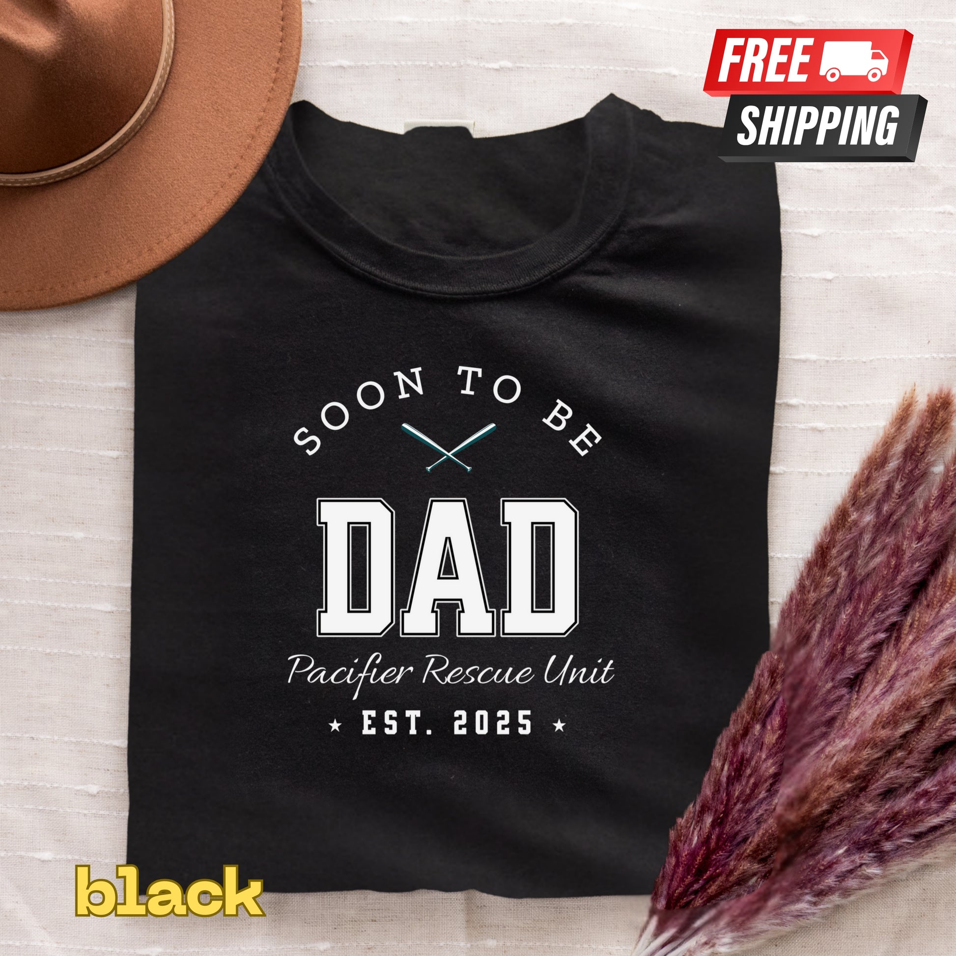Black t-shirt with 'Soon to be Dad' text on a textured surface with a hat and dried plants.