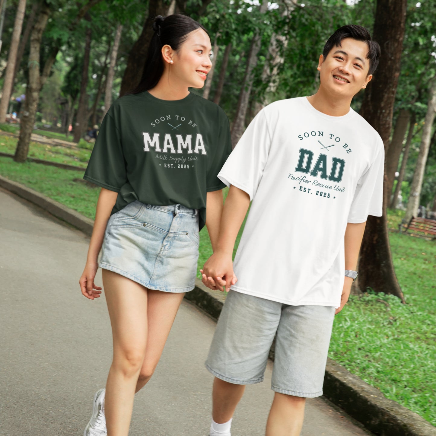 Two people holding hands, wearing 'Soon to be Mama' and 'Soon to be Dad' shirts in a park.