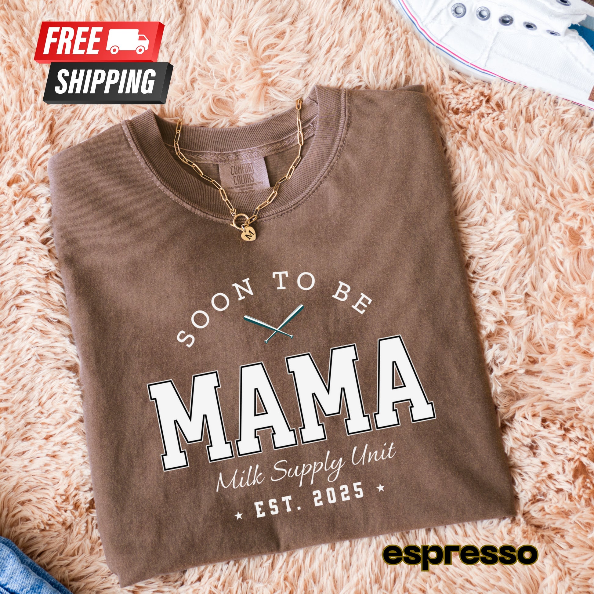 Folded brown sweatshirt with 'Soon to be Mama' text on a fluffy pink surface, featuring free shipping icon.