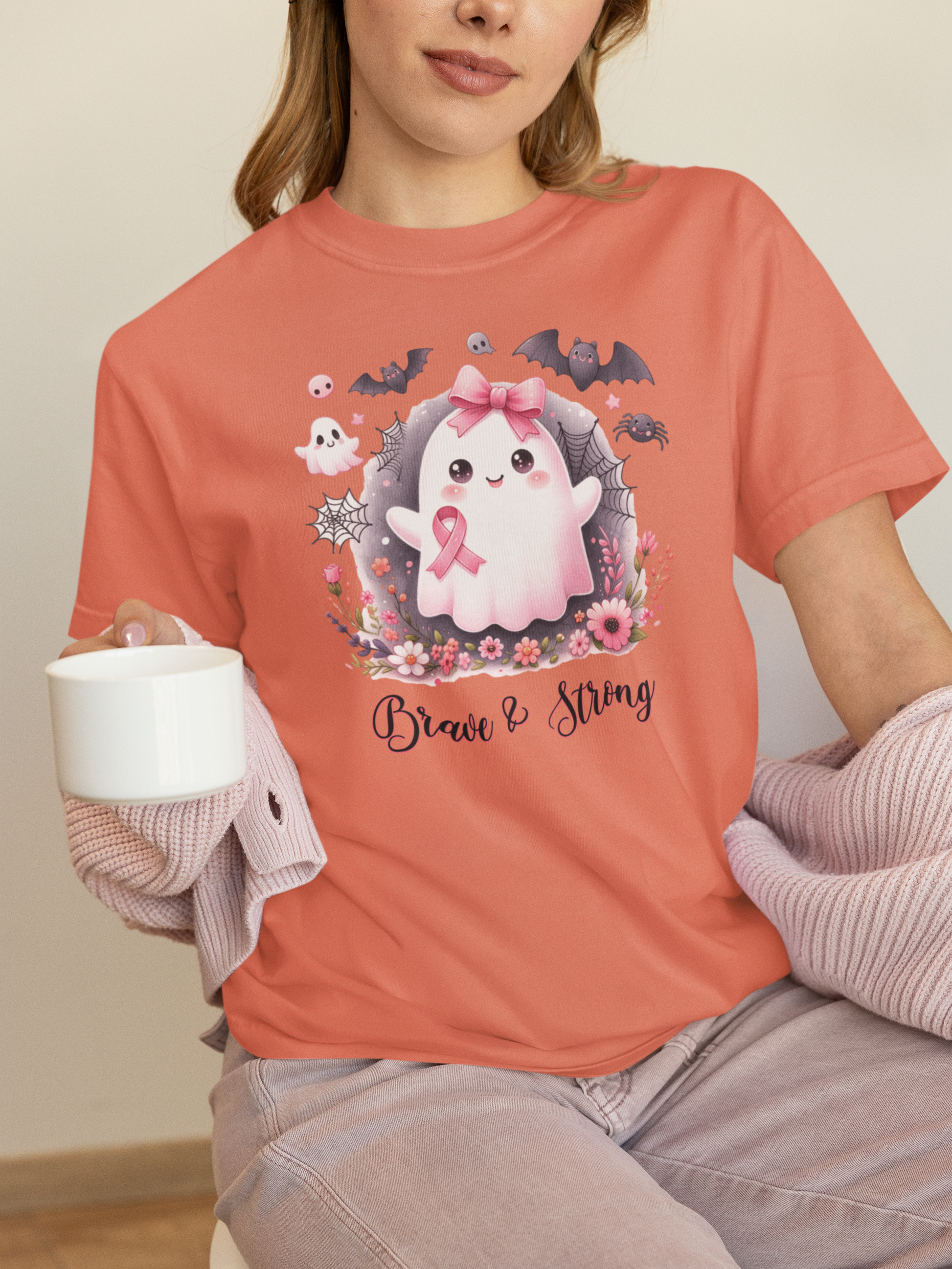 Person wearing a coral t-shirt with a ghost graphic and 'Brave & Strong' text, holding a white mug.