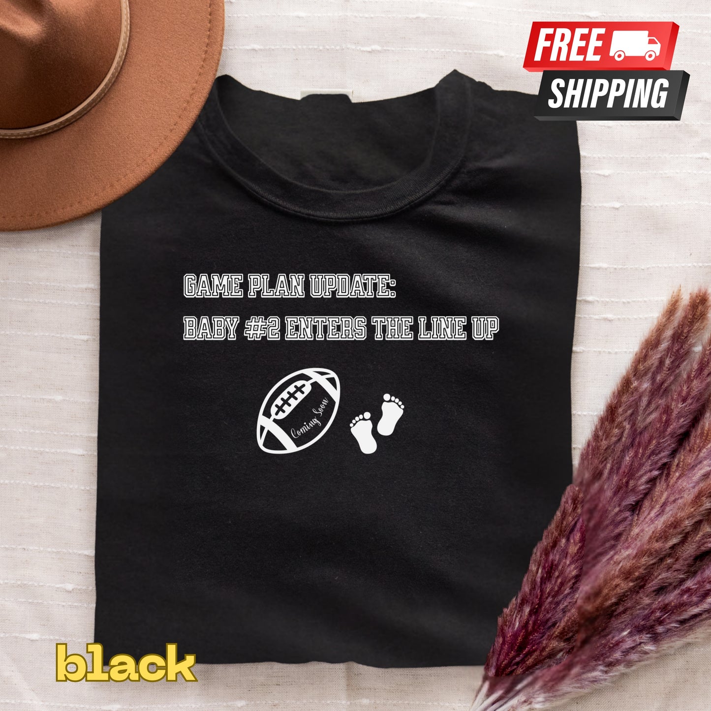 Black t-shirt with humorous text and graphics on a textured surface with a hat and dried plants.