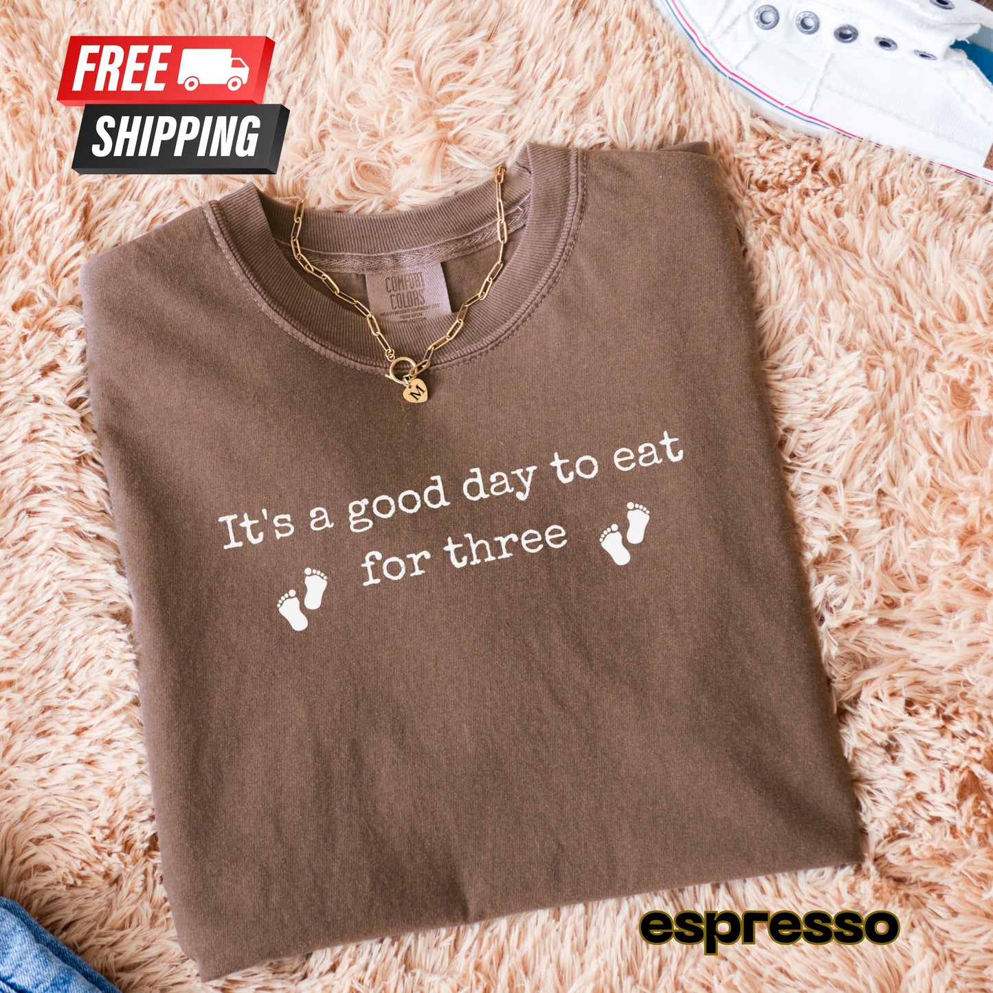 Folded brown t-shirt with text and baby footprints on a fluffy surface, featuring free shipping icon and espresso brand name.