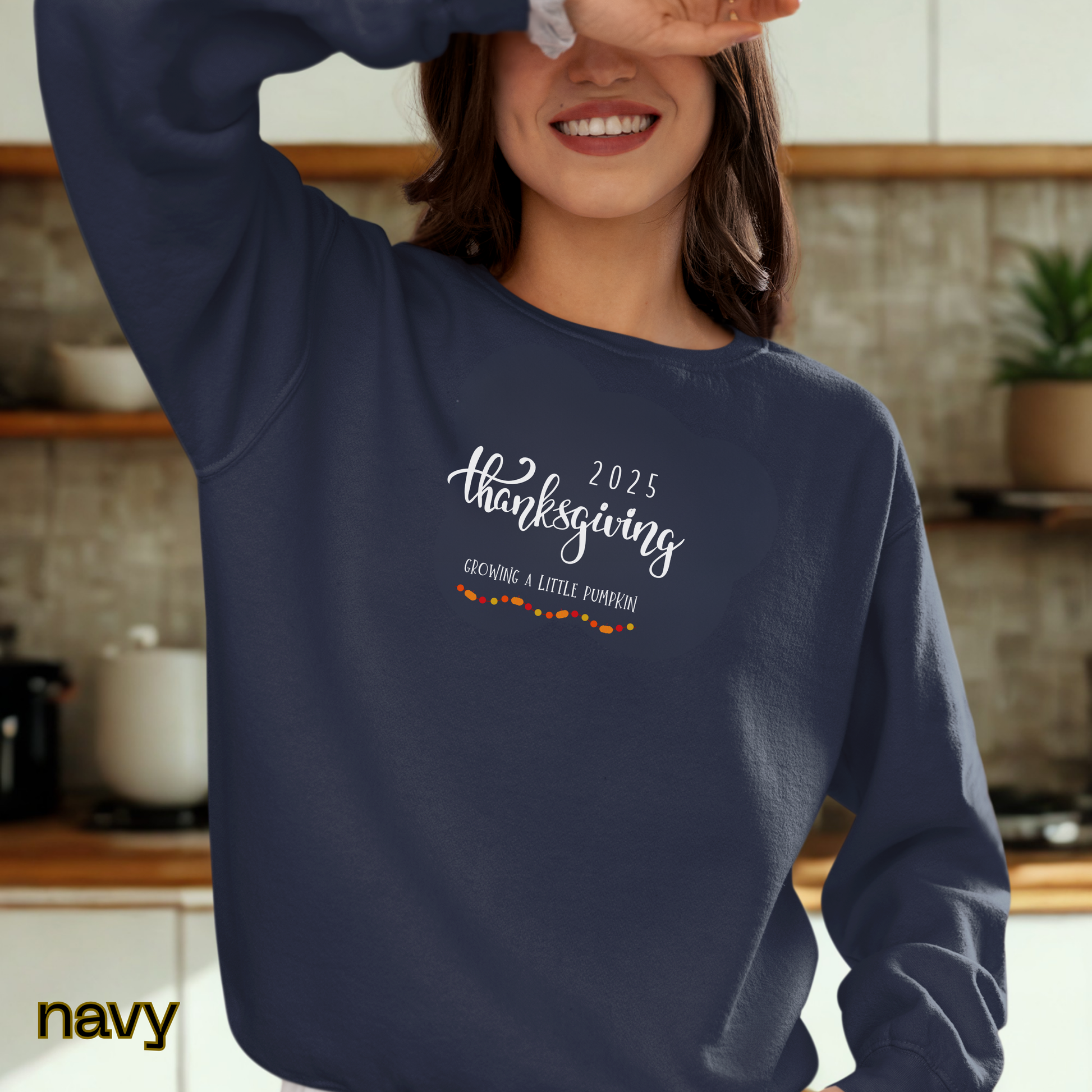 Person wearing a navy sweatshirt with '2025 Thanksgiving Growing a Little Pumpkin' text in a kitchen.