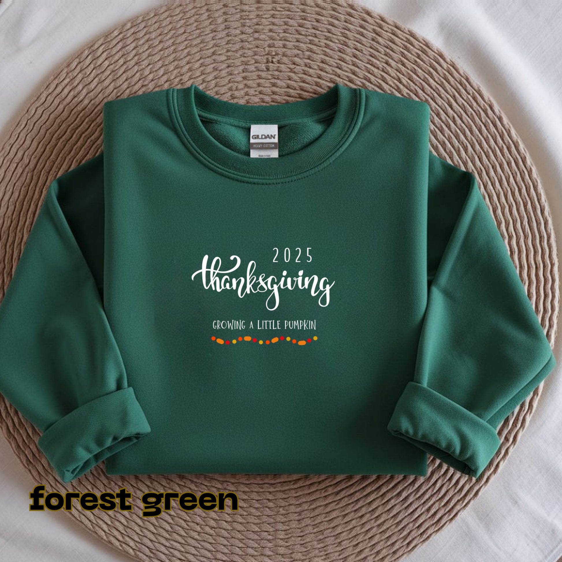 Folded green sweatshirt with '2025 Thanksgiving Growing a Little Pumpkin' text on a woven mat.