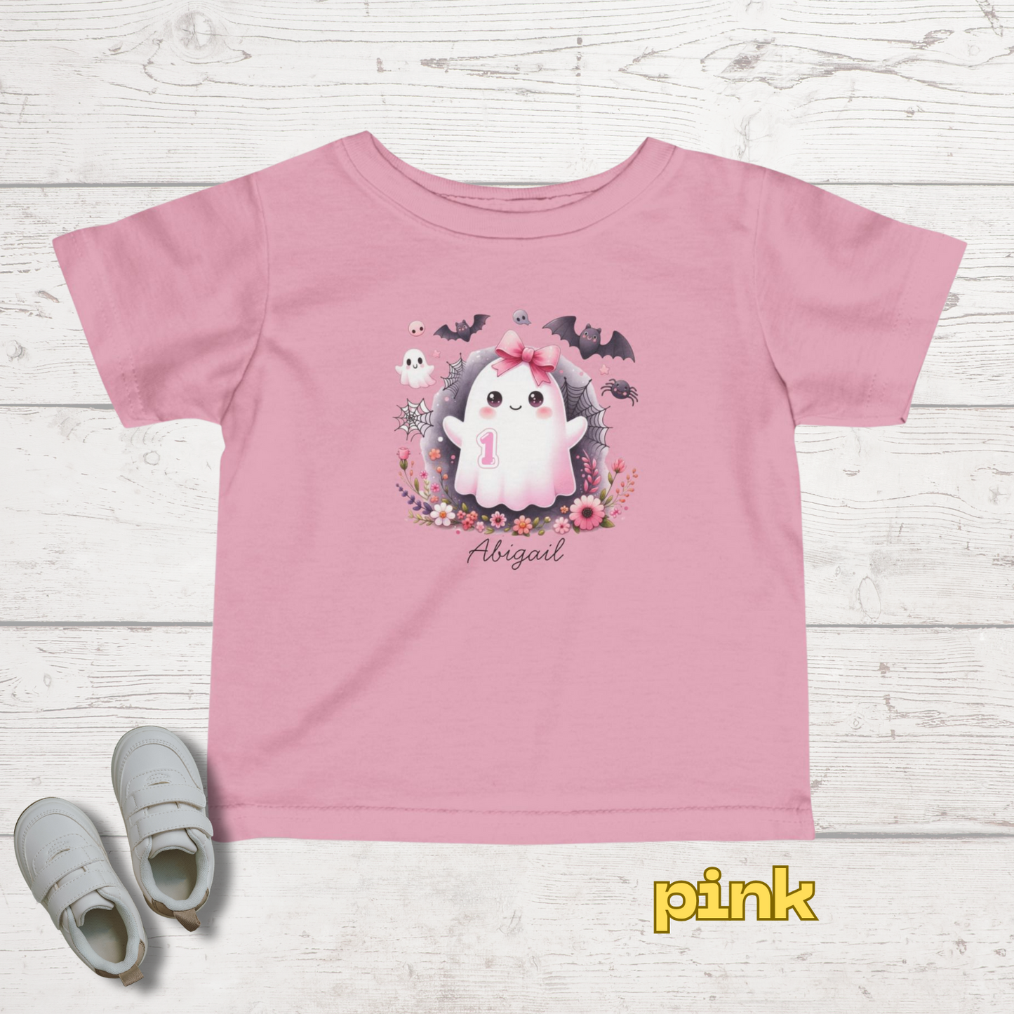 Pink t-shirt with a cartoon design and 'Abigail' text on a white wooden surface, with a pair of shoes beside it.